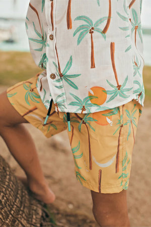A child stands barefoot outdoors in Pepita & Me—wearing a white shirt and Boys' Swim Trunks, both featuring green palm trees and orange suns on quick-dry fabric.