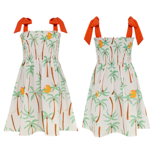 Two sleeveless Pepita & Me Girls' Waffle Dresses with orange shoulder ties feature a tropical palm tree and fruit print on white. The smocked top adds comfort and style—perfect for sunny days. Made in Colombia.