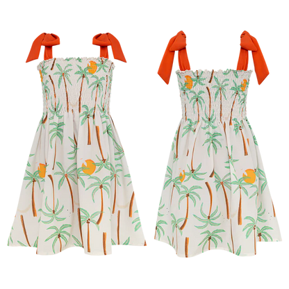 Two sleeveless Pepita & Me Girls' Waffle Dresses with orange shoulder ties feature a tropical palm tree and fruit print on white. The smocked top adds comfort and style—perfect for sunny days. Made in Colombia.