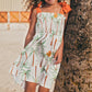 A young girl wears the Pepita & Me Girls' Waffle Dress—featuring a smocked top, palm tree prints, and orange shoulder ties—as she stands by a tree on a sandy beach.