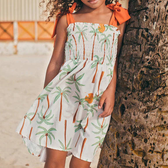 A young girl wears the Pepita & Me Girls' Waffle Dress—featuring a smocked top, palm tree prints, and orange shoulder ties—as she stands by a tree on a sandy beach.