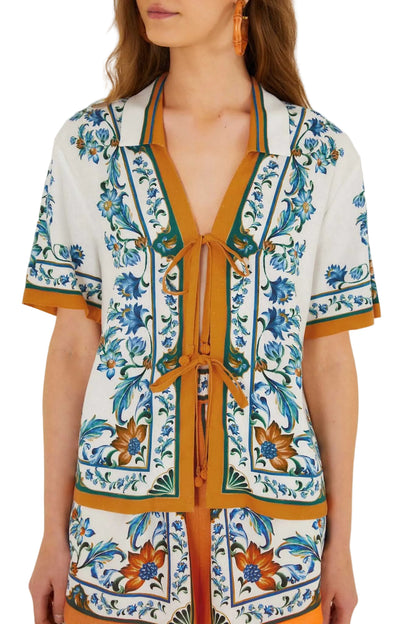 A woman wears the Farm Rio Flora Tile Off White Blouse, featuring short sleeves, a collar, front tie closure, and blue, green, and orange geometric floral patterns.