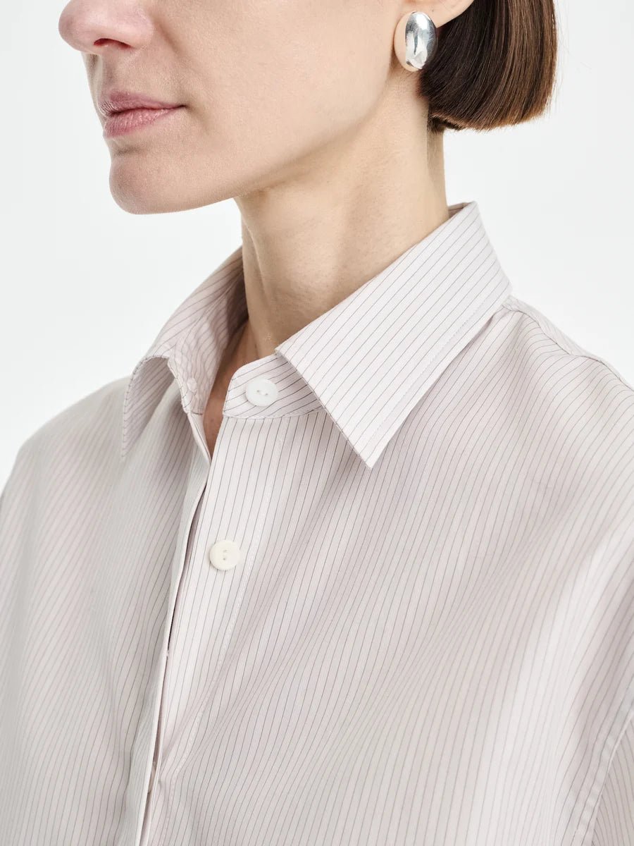 A woman shown from the shoulders up in side profile wears the Maria McManus Button Sleeve Crop Shirt, a light, pinstriped top made from Japanese Supima cotton, paired with silver earrings against a plain background.