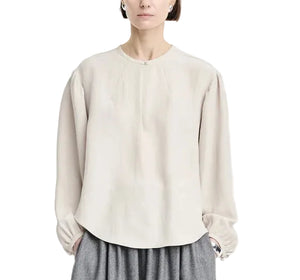 A person wearing the Maria McManus Pearl Button Blouse, a loose, long-sleeve beige top with gathered cuffs in matte sateen, paired with gray pants and standing against a white background.