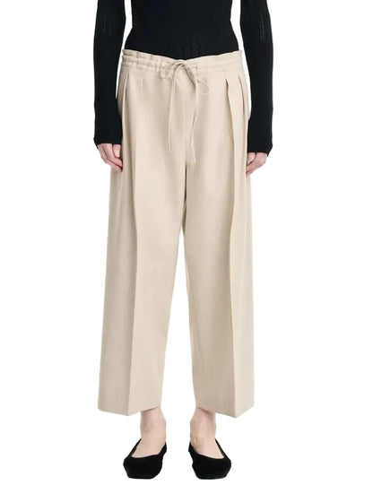 A person is wearing the Maria McManus Drawstring Trouser in beige wool-cashmere flannel with a wide-leg silhouette, styled with a black long-sleeve top and black flats, standing against a white background.