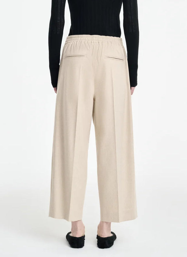 Model wears the Maria McManus Drawstring Trouser, featuring wide-leg, beige cropped loungewear style with an elastic waistband and two back welt pockets, paired with a black long-sleeve top and black shoes, standing against a white background.