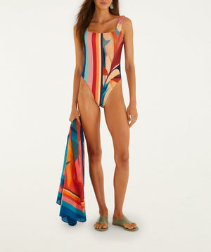 A woman wearing the Farm Rio Atlantic Rain Forest Light Pink Sarong holds it stylishly, pairing it with green slide sandals as she stands against a plain background.