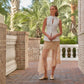 A woman stands on a brick patio, wearing a sleeveless white top, sheer beige skirt, and Sonya Metallic Loafers by Magnanni. Palm trees and greenery are in the background.