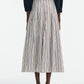 A person is seen from the back wearing the Lela Rose Striped Cotton Full Skirt—a cream pleated A-line with thin blue and brown stripes—paired with a dark top and brown sandals.