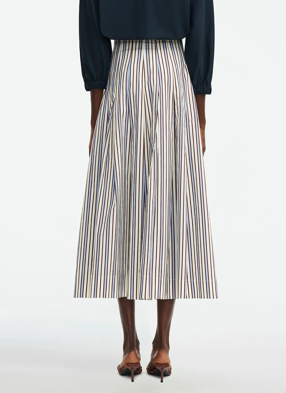 A person is seen from the back wearing the Lela Rose Striped Cotton Full Skirt—a cream pleated A-line with thin blue and brown stripes—paired with a dark top and brown sandals.
