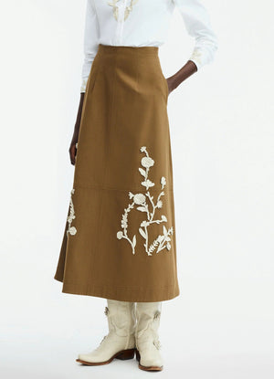 A person in the Lela Rose Leather Floral Embroidered Rustic Skirt, crisp white shirt, and white cowboy boots stands against a plain background.