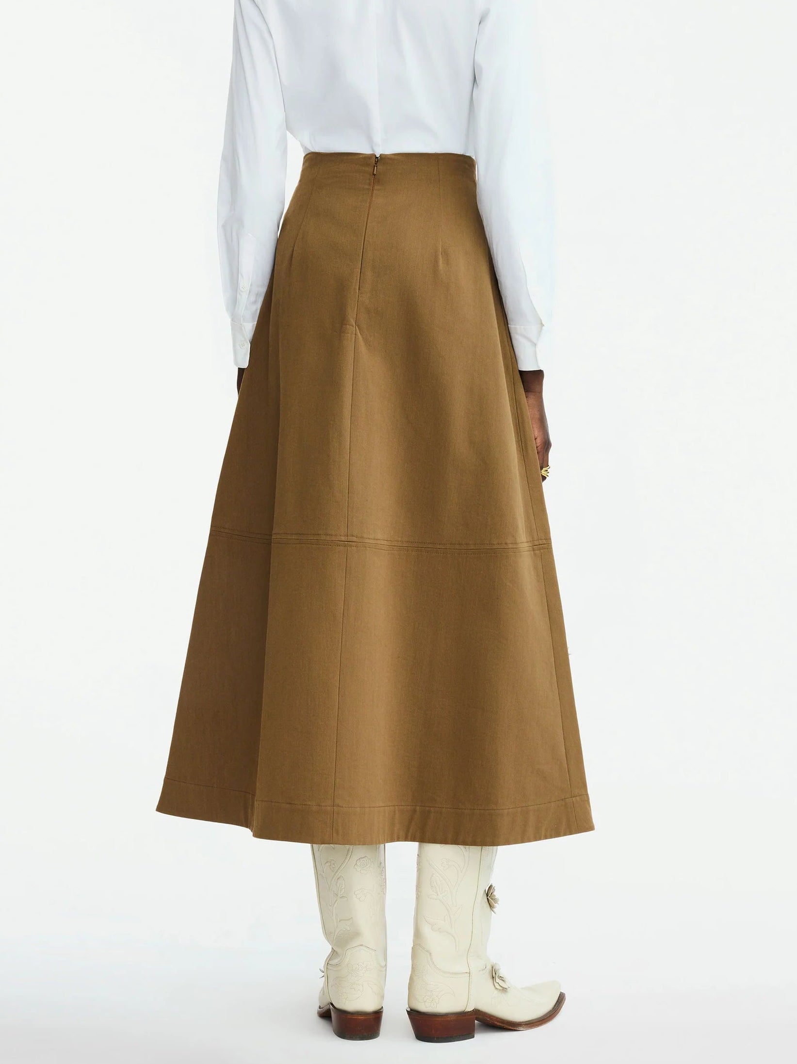 A person is seen from the back wearing a white long-sleeve shirt, the Lela Rose Leather Floral Embroidered Rustic Skirt in brown, and white cowboy boots, set against a plain background.