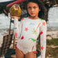 A young girl in the Pepita & Me Baby Girls' and Girls' Knot LS Rashguard enjoys sun protection and style by the beach, holding a coconut with palm debris and painted tires in the background.