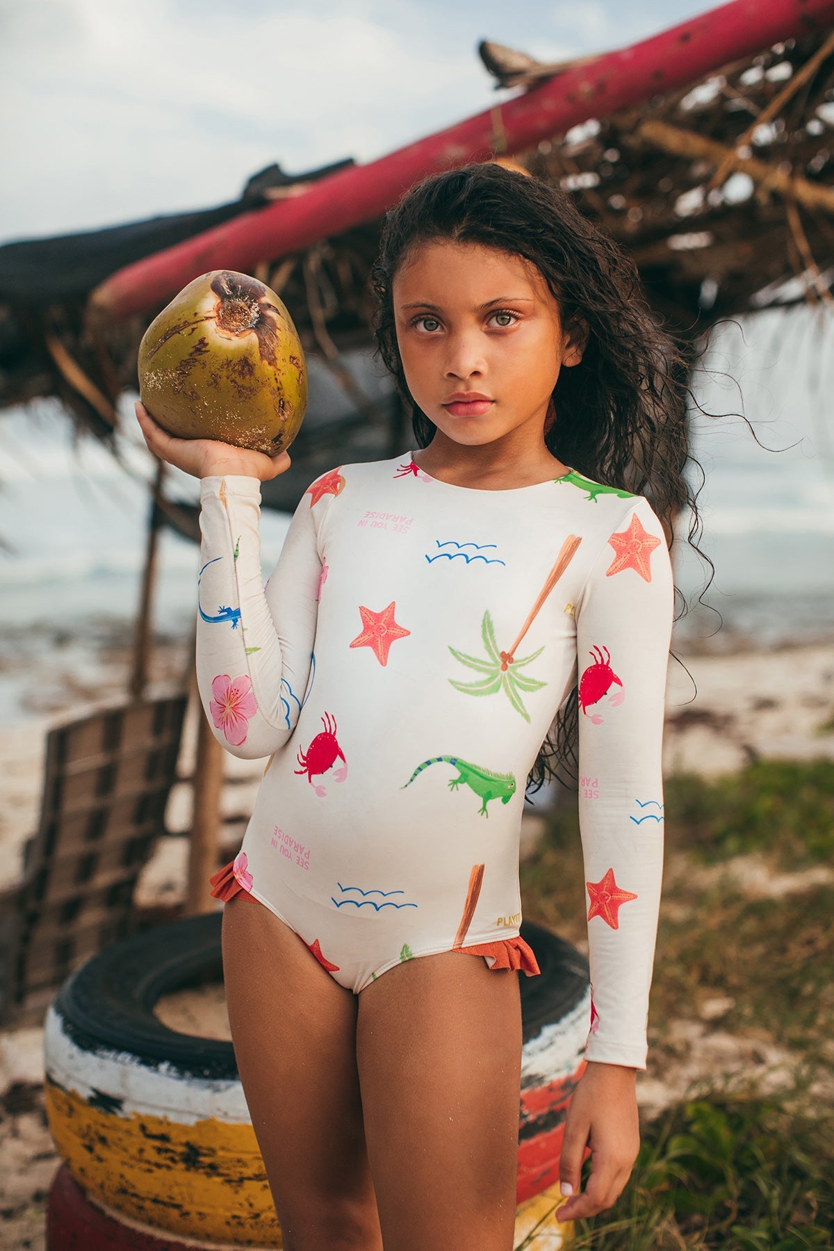 A young girl in the Pepita & Me Baby Girls' and Girls' Knot LS Rashguard enjoys sun protection and style by the beach, holding a coconut with palm debris and painted tires in the background.