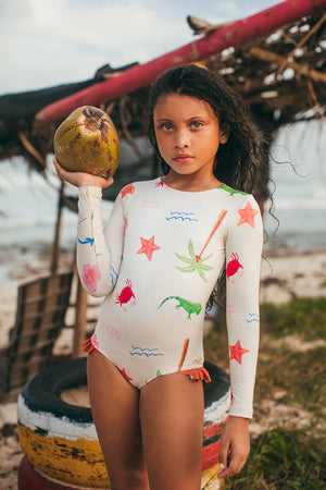 A young girl in the Pepita & Me Baby Girls' and Girls' Knot LS Rashguard enjoys sun protection and style by the beach, holding a coconut with palm debris and painted tires in the background.