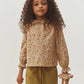 A young girl with curly hair wears the Bonpoint Girls’ Melusina Blouse in Liberty fabric, featuring long sleeves, ruffled cuffs, and a delicate floral print, paired with olive green pants as she stands against a light background.