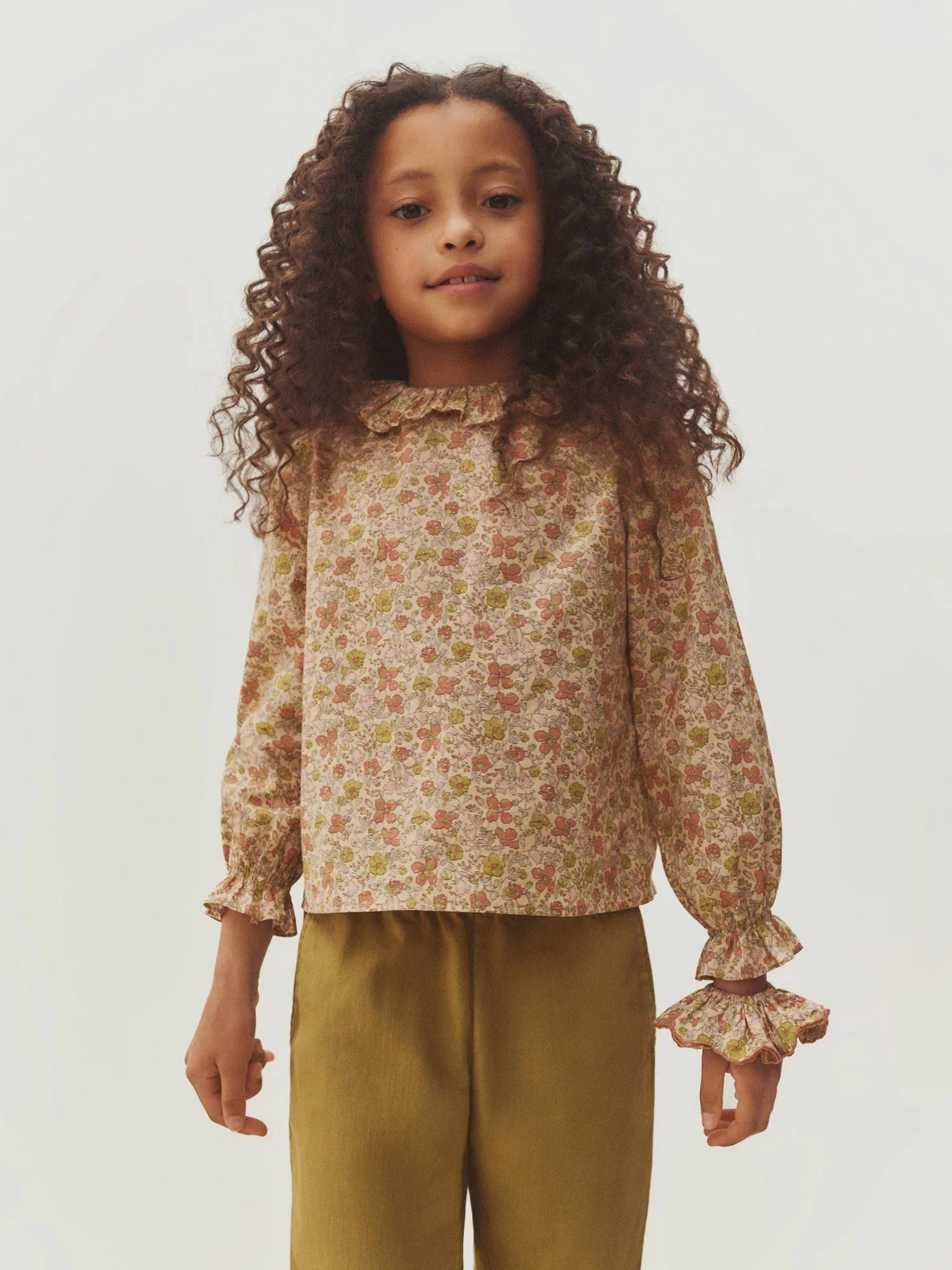 A young girl with curly hair wears the Bonpoint Girls’ Melusina Blouse in Liberty fabric, featuring long sleeves, ruffled cuffs, and a delicate floral print, paired with olive green pants as she stands against a light background.