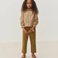 A young girl with long curly hair stands facing forward against a plain white background, wearing a floral blouse with ruffled sleeves, Bonpoint Girls’ July Pants crafted from organic cotton in a mustard color, and pink shoes.