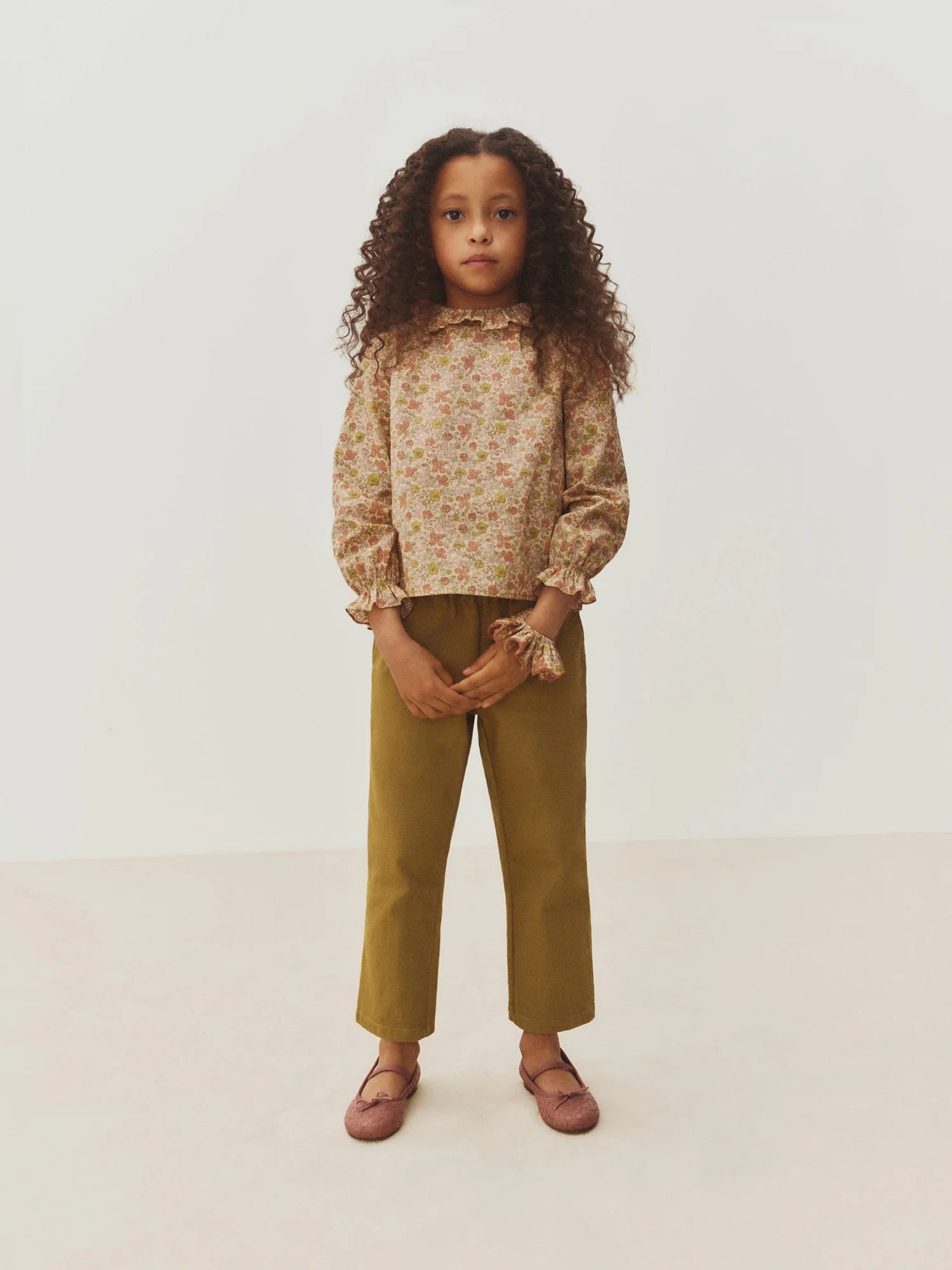 A young girl with long curly hair stands facing forward against a plain white background, wearing a floral blouse with ruffled sleeves, Bonpoint Girls’ July Pants crafted from organic cotton in a mustard color, and pink shoes.
