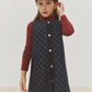 A young child in the Bonpoint Girls’ Jette Cherry Checkerboard-Print Dress, made from organic cotton denim, stands against a plain background wearing a red turtleneck and cap, with a pencil tucked behind their ear.