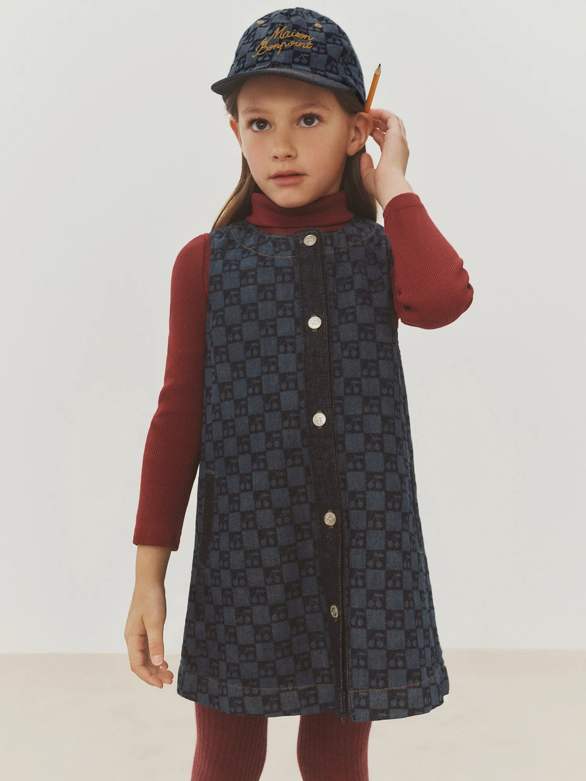 A young child in the Bonpoint Girls’ Jette Cherry Checkerboard-Print Dress, made from organic cotton denim, stands against a plain background wearing a red turtleneck and cap, with a pencil tucked behind their ear.