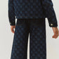 A person is seen from the back wearing Bonpoint’s Girls’ Minalie Cherry Checkerboard-Print Denim Trousers featuring a blue checkerboard pattern and an adjustable high waist, paired with a matching denim jacket.