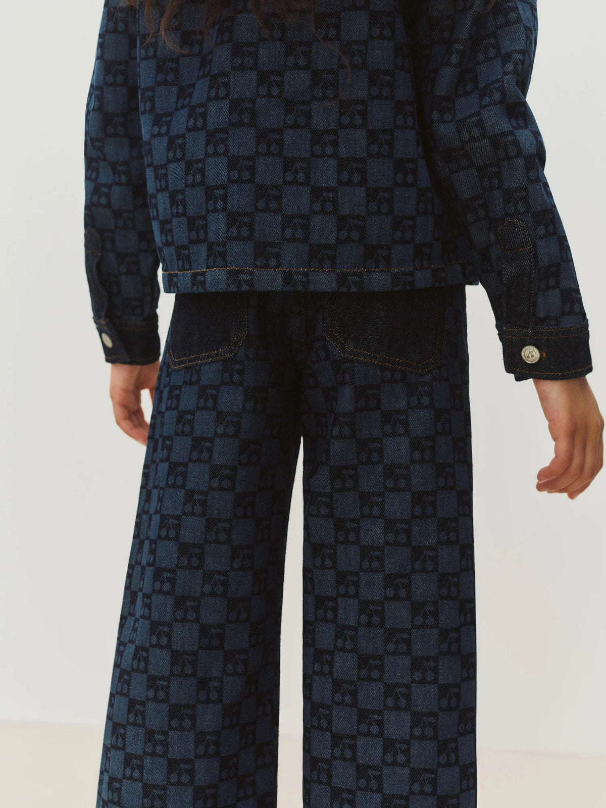 A person is seen from the back wearing Bonpoint’s Girls’ Minalie Cherry Checkerboard-Print Denim Trousers featuring a blue checkerboard pattern and an adjustable high waist, paired with a matching denim jacket.