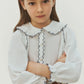 A young girl with straight brown hair wears the Bonpoint Girls’ Maneke Blouse with Peter Pan Collar Embroidery, an organic cotton blouse featuring a vintage collar and ruffled details. She pairs it with a patterned skirt and stands with her arms crossed, looking at the camera.