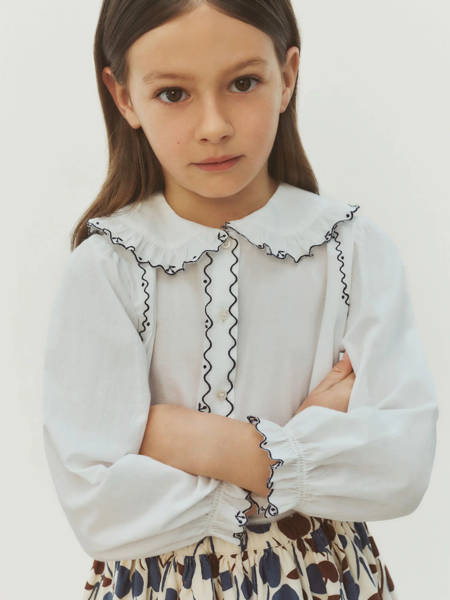 A young girl with straight brown hair wears the Bonpoint Girls’ Maneke Blouse with Peter Pan Collar Embroidery, an organic cotton blouse featuring a vintage collar and ruffled details. She pairs it with a patterned skirt and stands with her arms crossed, looking at the camera.