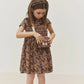 A young girl stands against a plain background, gazing into a small purse adorned with a pink cherry charm. She wears the Bonpoint Girls’ Duchesse Dress, crafted from organic cotton Liberty fabric in an elegant floral print, paired with a matching headband for a charming look.