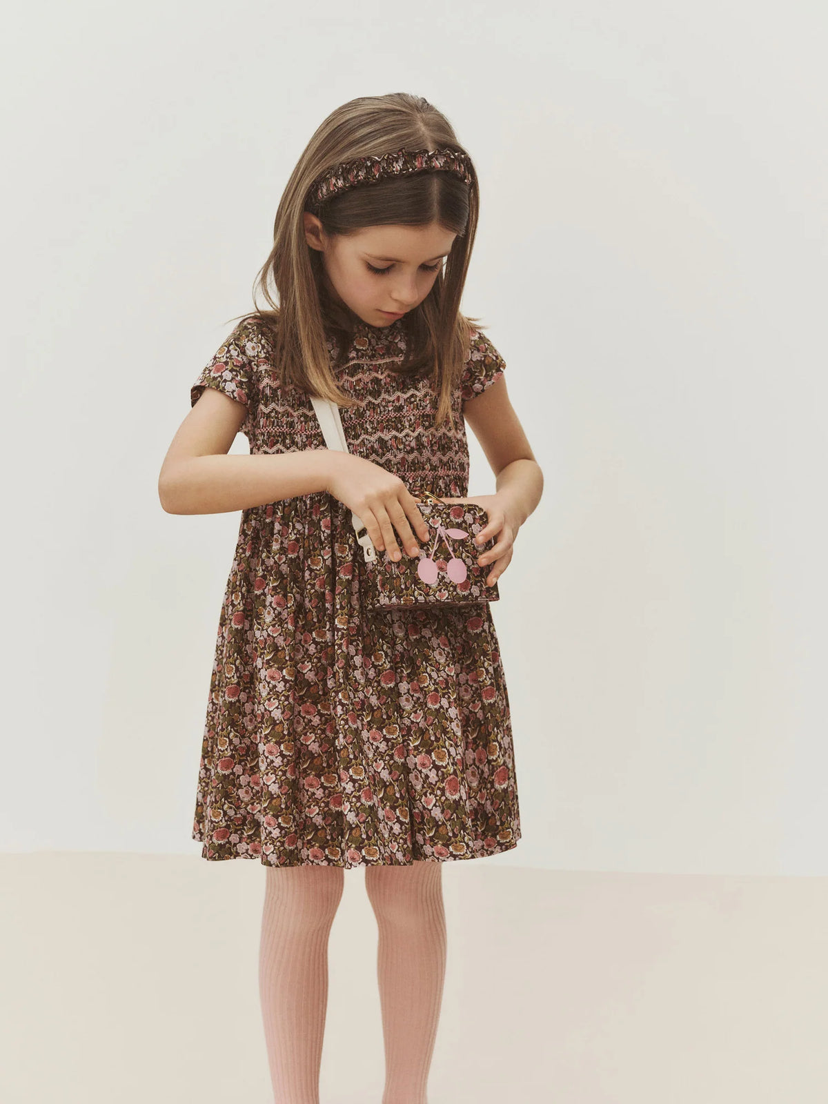 A young girl stands against a plain background, gazing into a small purse adorned with a pink cherry charm. She wears the Bonpoint Girls’ Duchesse Dress, crafted from organic cotton Liberty fabric in an elegant floral print, paired with a matching headband for a charming look.