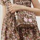 A child in a floral dress holds the Bonpoint Girls’ Aimane Travel Bag, a matching floral purse with pink cherries on the front, crafted from soft Liberty fabric. The purse features a white strap and is perfect as a suitcase bag for girls.