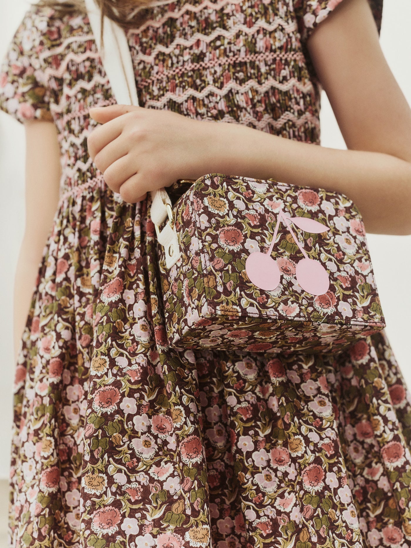 A child in a floral dress holds the Bonpoint Girls’ Aimane Travel Bag, a matching floral purse with pink cherries on the front, crafted from soft Liberty fabric. The purse features a white strap and is perfect as a suitcase bag for girls.