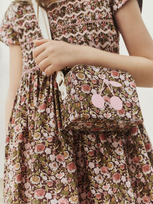 A child in a floral dress holds the Bonpoint Girls’ Aimane Travel Bag, a matching floral purse with pink cherries on the front, crafted from soft Liberty fabric. The purse features a white strap and is perfect as a suitcase bag for girls.