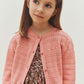 A young girl wears the Bonpoint Girls’ Mia Cardigan in pink over a floral dress, standing against a plain light background while looking slightly to the side.