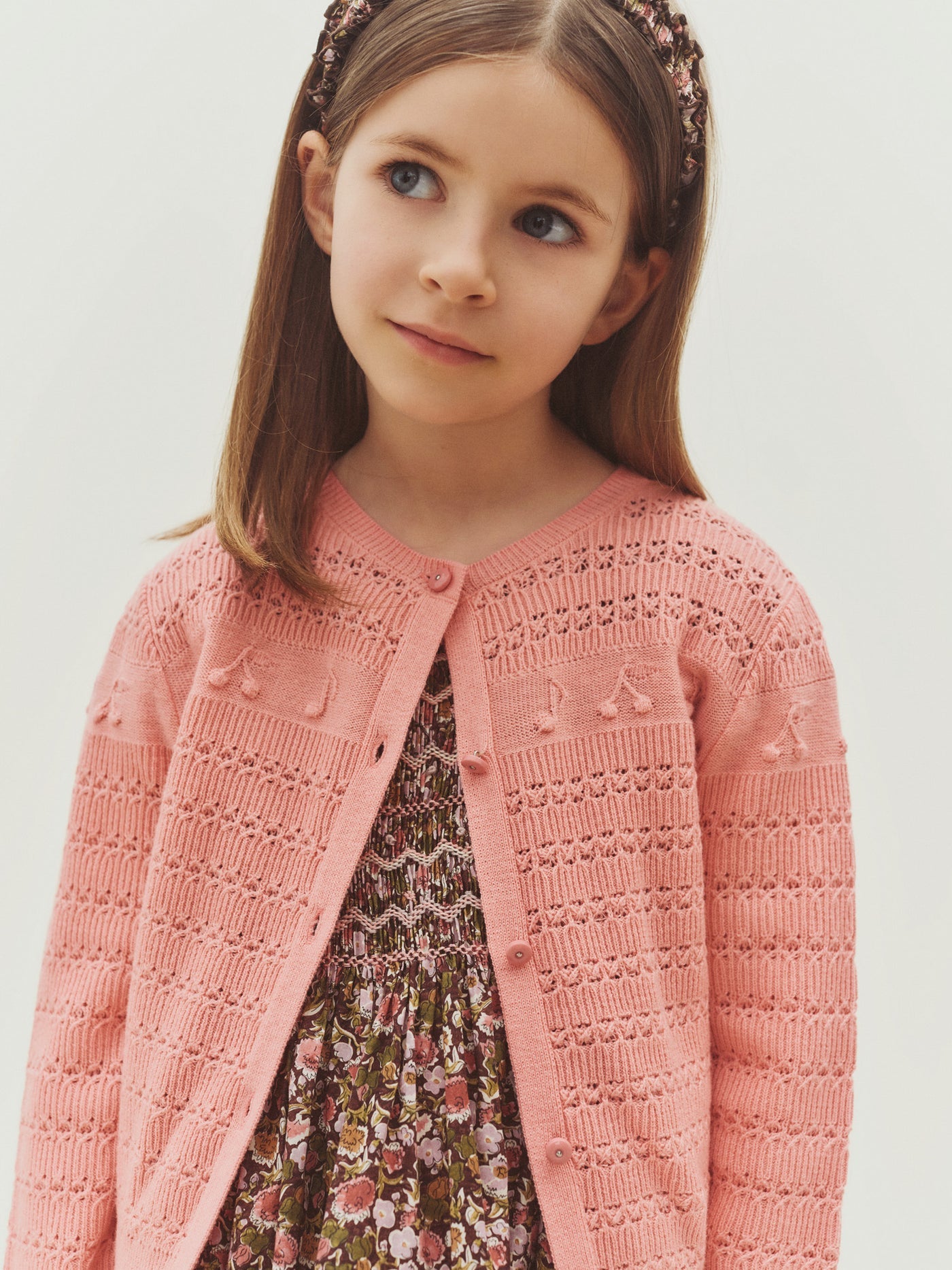 A young girl wears the Bonpoint Girls’ Mia Cardigan in pink over a floral dress, standing against a plain light background while looking slightly to the side.