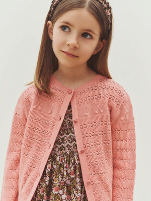 A young girl wears the Bonpoint Girls’ Mia Cardigan in pink over a floral dress, standing against a plain light background while looking slightly to the side.