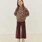 A young girl stands with her hands in her pockets, wearing the Bonpoint Girls’ Petale Blouse crafted from Liberty fabric with hand-embroidered smocking, paired with burgundy wide-leg pants, metallic boots, and a hair clip, set against a plain light background.