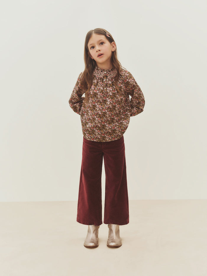 A young girl stands with her hands in her pockets, wearing the Bonpoint Girls’ Petale Blouse crafted from Liberty fabric with hand-embroidered smocking, paired with burgundy wide-leg pants, metallic boots, and a hair clip, set against a plain light background.
