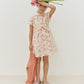 A young girl wears the Bonpoint Girls’ Gisele Dress, featuring a delicate floral print, as she holds two pink flowers to her forehead and carries a pink sweater, standing against a plain white background.