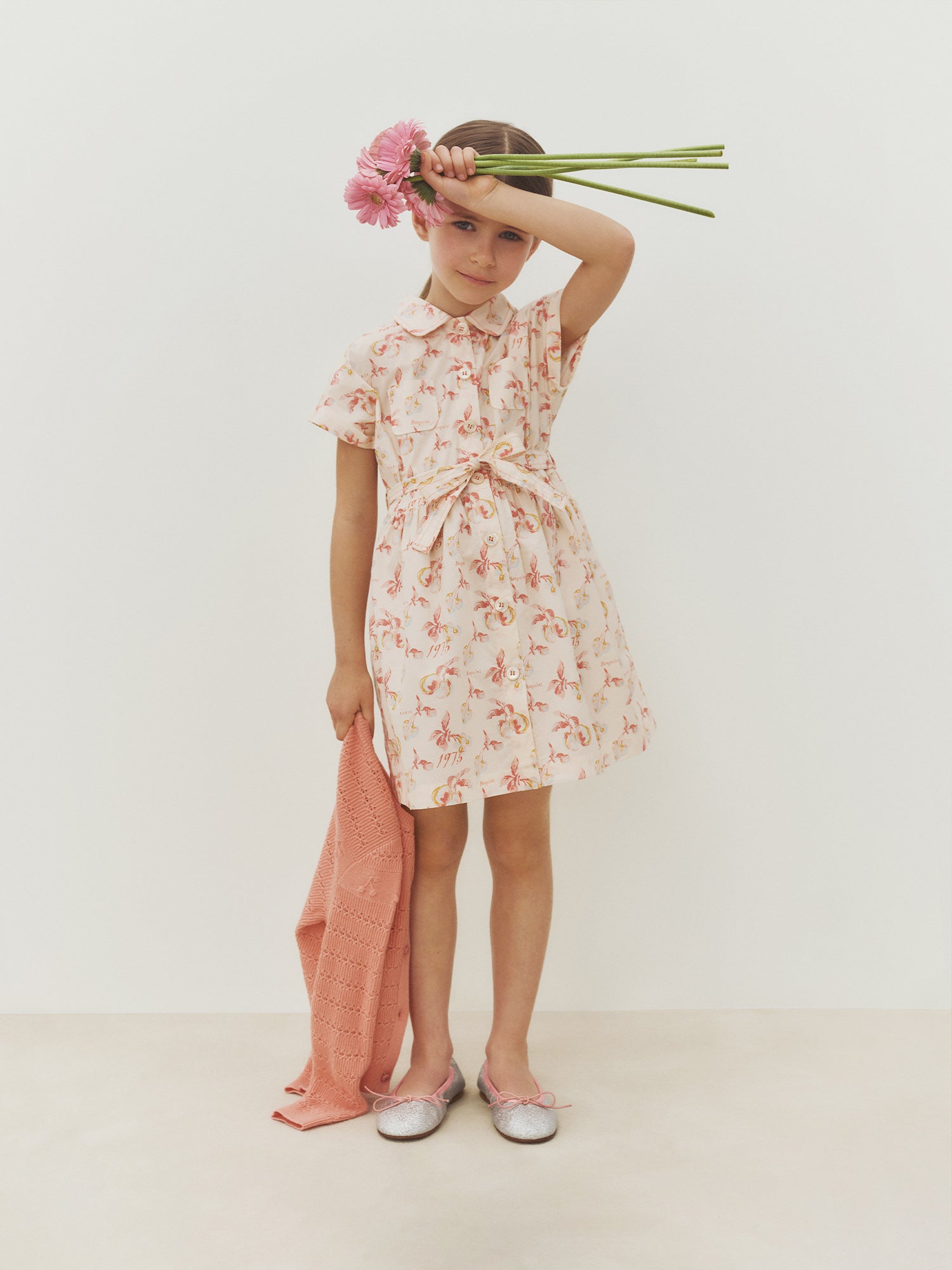 A young girl wears the Bonpoint Girls’ Gisele Dress, featuring a delicate floral print, as she holds two pink flowers to her forehead and carries a pink sweater, standing against a plain white background.