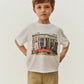 A young boy stands against a plain background wearing the Boys’ Eidan T-Shirt by Bonpoint, crafted from organic cotton and featuring a car and building graphic, paired with beige pants.