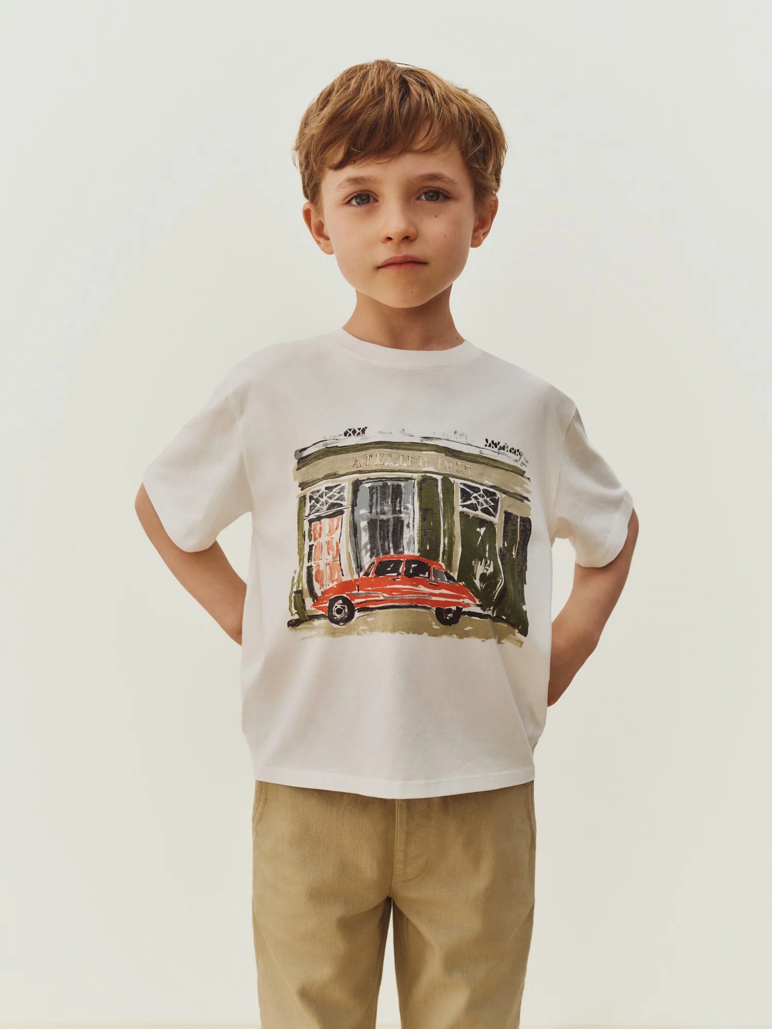 A young boy stands against a plain background wearing the Boys’ Eidan T-Shirt by Bonpoint, crafted from organic cotton and featuring a car and building graphic, paired with beige pants.