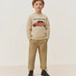 A young boy stands facing forward against a plain light background, wearing a cream sweater with a car graphic and text, Bonpoint Boys’ Maurice Straight Pants in tan with an elastic waist, and black boots.
