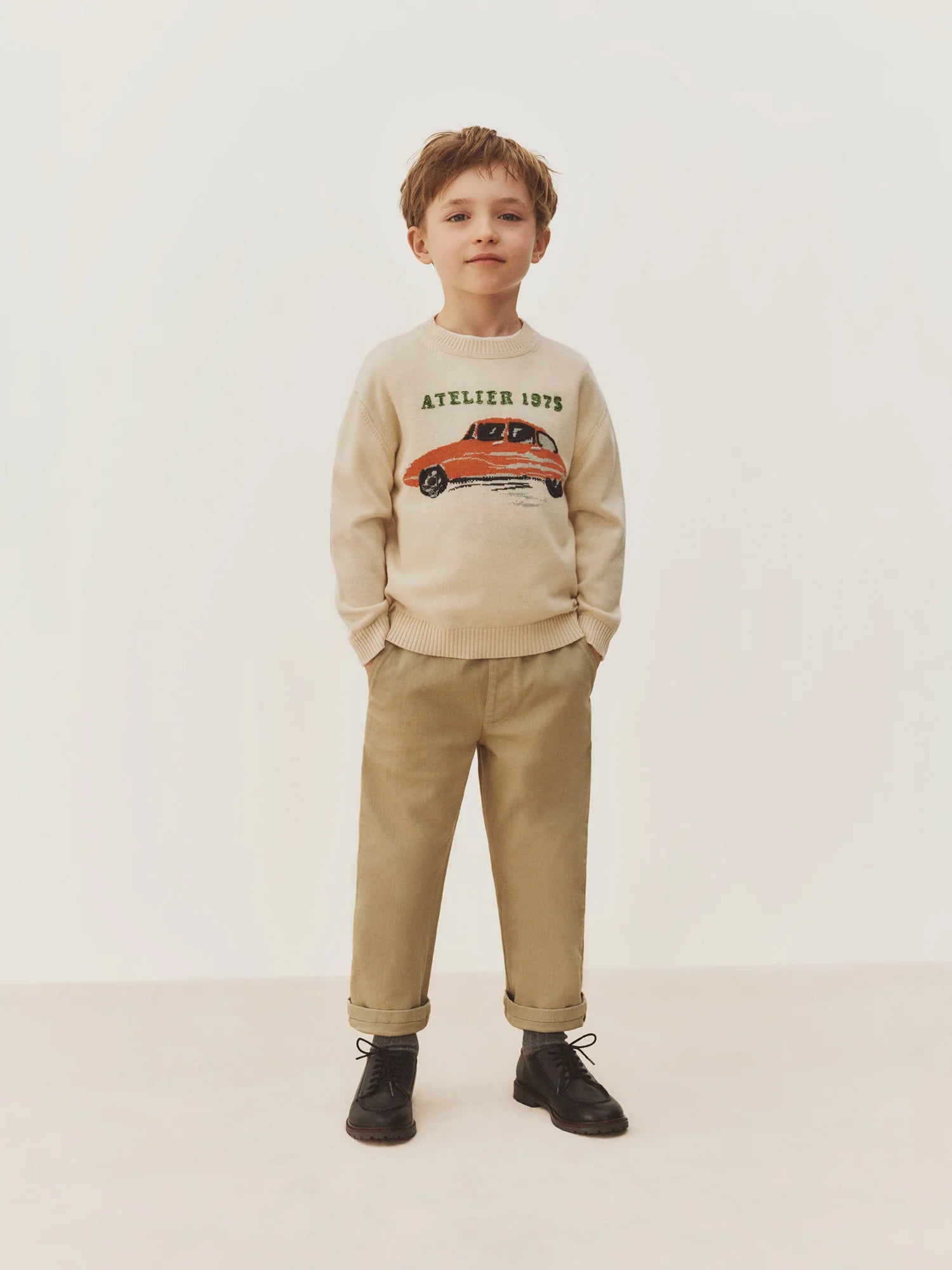 A young boy stands facing forward against a plain light background, wearing a cream sweater with a car graphic and text, Bonpoint Boys’ Maurice Straight Pants in tan with an elastic waist, and black boots.