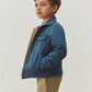 A young child with short brown hair wears the Bonpoint Boys’ Marvin Reversible Jacket with Embroidery on the Back, paired with tan pants, standing against a plain light background and looking slightly upward.