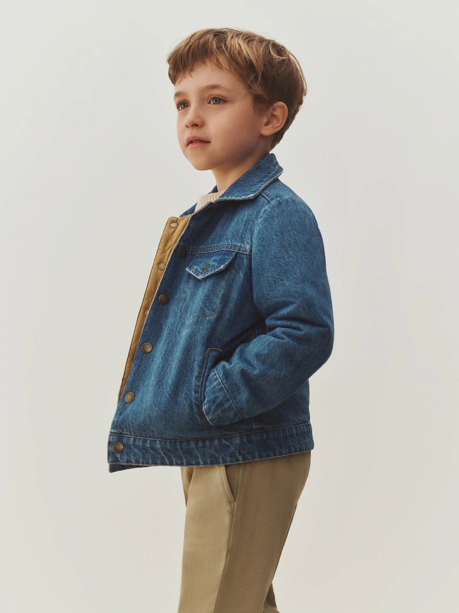 A young child with short brown hair wears the Bonpoint Boys’ Marvin Reversible Jacket with Embroidery on the Back, paired with tan pants, standing against a plain light background and looking slightly upward.