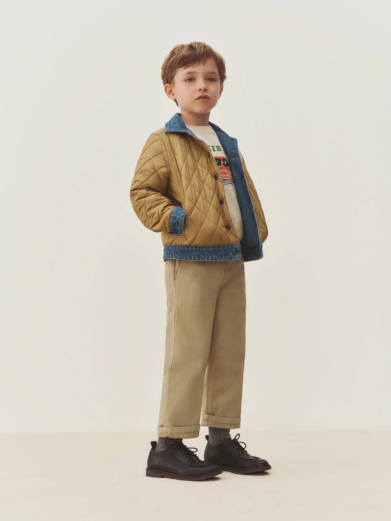 A young child stands facing forward against a plain off-white background, wearing the Bonpoint Boys’ Marvin Reversible Jacket with Embroidery on the Back, featuring denim trim, paired with beige pants, black shoes, and a graphic T-shirt.