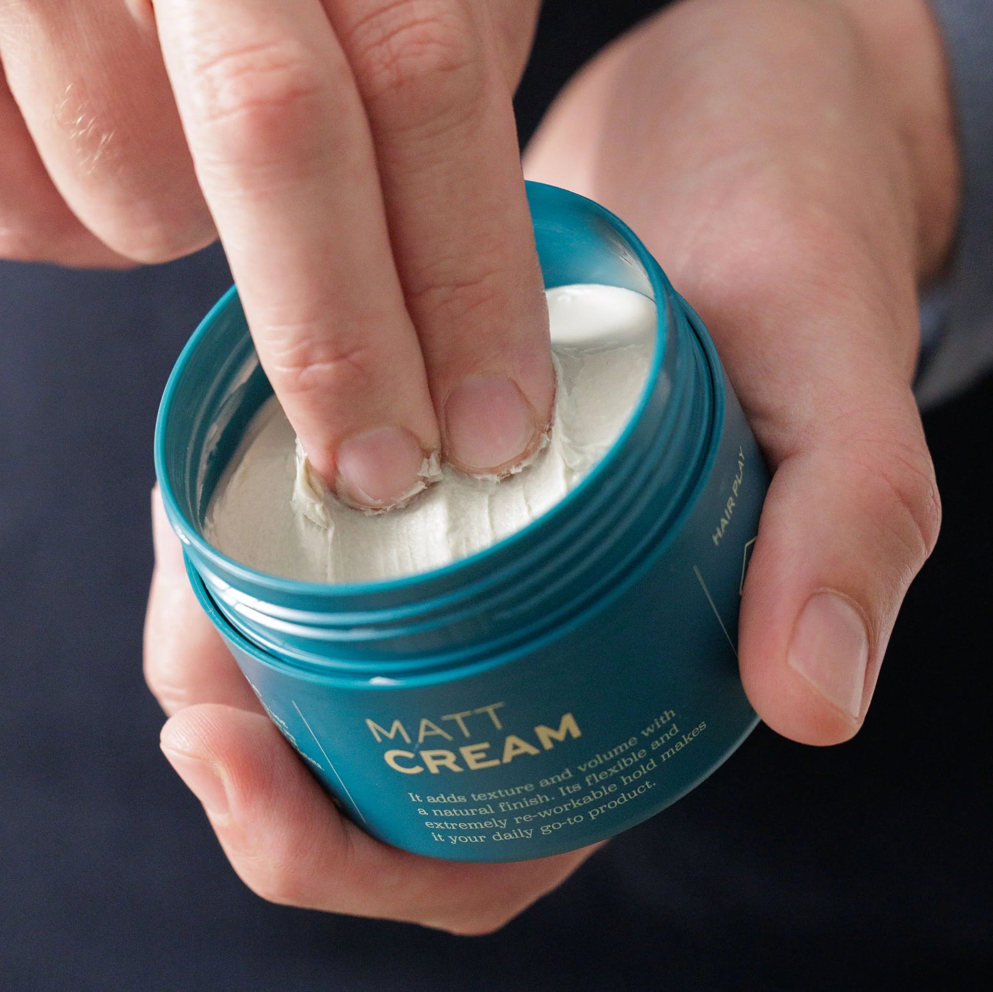 A person scoops Barberino's Matt Cream, a white hair styling cream with medium hold and anti-frizz benefits, from a blue container using their fingers.