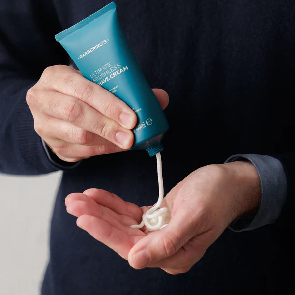 A person squeezes Barberino's Ultimate Brushless Shave Cream from a teal tube onto their palm, beginning their grooming routine for a smooth shave.